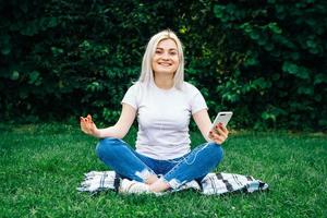 la donna con le cuffie e lo smartphone in mano siede in posa meditativa ascoltando musica sull'erba verde