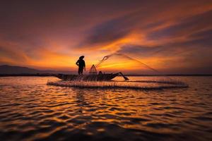 sagoma di pescatore asiatico su barca di legno in azione gettando una rete per catturare pesci d'acqua dolce nel fiume naturale al mattino presto prima dell'alba foto