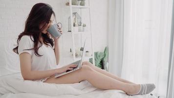 ritratto di bella donna asiatica attraente che utilizza computer o laptop in possesso di una calda tazza di caffè o tè mentre è sdraiata sul letto quando si rilassa nella sua camera da letto a casa. concetto di stile di vita delle donne a casa. foto