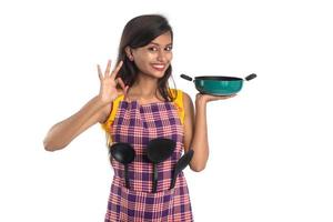 Giovane donna indiana tenendo arnese da cucina cucchiaio, stapula, mestolo e padella, ecc. su sfondo bianco foto