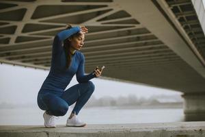 giovane donna in tuta da ginnastica blu che monitora i suoi progressi di allenamento sull'app fitness vicino al fiume al mattino d'autunno