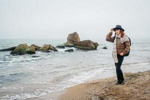 donna con cappello e sciarpa con uno zaino sulla costa sullo sfondo delle rocce contro il bellissimo mare foto