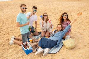 un' gruppo di cinque amici rilassa su il sabbioso spiaggia, Tenere bevande e giocoso oggetti di scena, godendo un' spensierato giorno insieme sotto il sole, condivisione risata e estate vibrazioni. foto