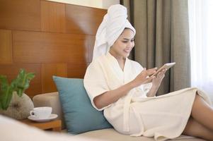 una donna rilassata che indossa un accappatoio bianco e un asciugamano che beve un caffè mentre usa il tablet al mattino foto