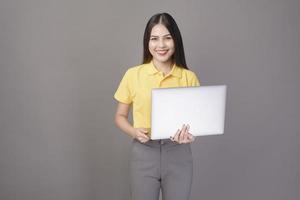 giovane bella donna sicura di sé che indossa una camicia gialla sta tenendo un laptop su sfondo grigio studio foto