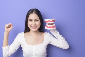 la giovane bella donna sorridente sta tenendo i denti della protesi di plastica sopra lo studio porpora isolato del fondo foto