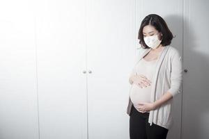 la bella donna incinta asiatica indossa una maschera protettiva nella sua casa, concetto di protezione del coronavirus foto