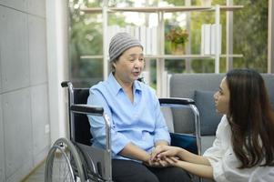 donna malata di cancro che indossa una sciarpa per la testa seduta su sedia a rotelle che parla con sua figlia di supporto all'interno, concetto di salute e assicurazione.