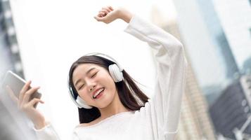ragazza asiatica sorridente felice che indossa le cuffie che ascolta e si muove alla musica contro la priorità bassa dell'edificio della città foto