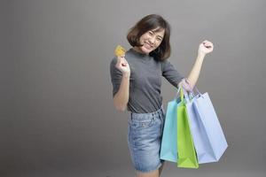 Ritratto di giovane bella donna asiatica in possesso di carta di credito e shopping bag colorato isolato su sfondo grigio studio foto