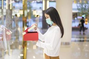 donna che indossa una maschera protettiva con in mano una confezione regalo nel centro commerciale, lo shopping sotto la pandemia covid-19, il ringraziamento e il concetto di natale.