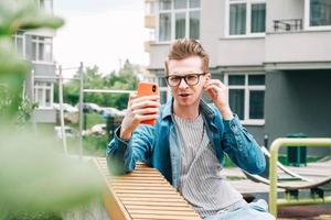 ritratto uomo in camicia e occhiali che utilizza comunica su uno smartphone seduto su una panchina su uno sfondo di case foto