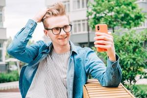 l'uomo in camicia e occhiali che usa comunica su uno smartphone seduto su una panchina su uno sfondo di case foto