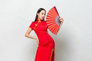 bella donna asiatica in abito rosso in stile orientale qipao che tiene il ventilatore con un'altra mano in gesto akimbo su sfondo grigio chiaro studio isolato foto