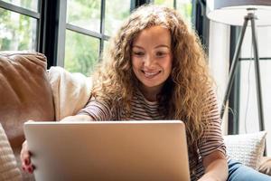 donna caucasica piuttosto sorridente che effettua una videochiamata con il computer portatile sul divano nel soggiorno di casa durante il giorno foto