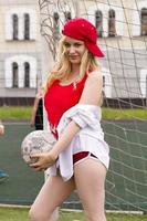 la bionda in forma rossa con una palla al cancello del campo di calcio. foto