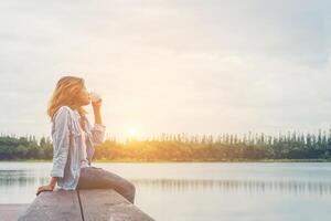 giovane bella donna hipster che tiene una tazza di caffè seduta sul lago rilassante con l'aria fresca e l'alba al mattino. foto