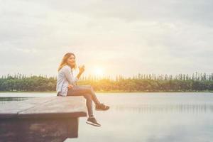 giovane bella donna hipster che tiene una tazza di caffè seduta sul lago rilassante con l'aria fresca e l'alba al mattino. foto