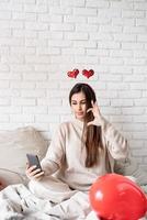 giovane donna divertente seduta nel letto per celebrare il giorno di San Valentino in chat utilizzando il telefono cellulare