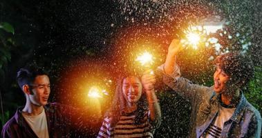 gruppo asiatico di amici che hanno barbecue in giardino all'aperto ridendo con bevande alcoliche di birra e mostrando un gruppo di amici che si divertono con le stelle filanti la notte foto