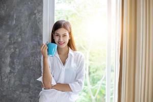 donne asiatiche che bevono caffè e si svegliano nel suo letto completamente riposate e aprono le tende al mattino per prendere aria fresca al sole foto