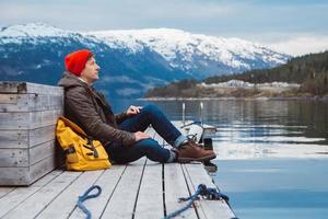uomo con uno zaino giallo che indossa un cappello rosso seduto sul molo di legno sullo sfondo della montagna e del lago foto
