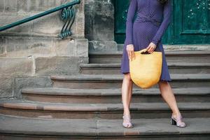 donna che indossa un abito a pois viola, con in mano una borsa gialla lavorata a maglia in piedi sulle scale in posa in una strada della città. copia, spazio vuoto per il testo
