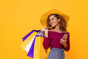 donna di razza mista sorridente alla moda con borse della spesa colorate e telefono cellulare isolato su sfondo giallo per conept vendita estiva foto