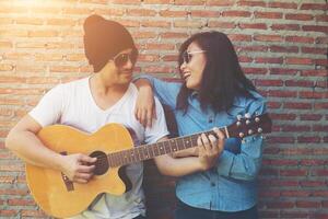 la coppia di hipster si abbraccia, distoglie lo sguardo e sorride mentre è in piedi all'aperto a suonare la chitarra contro un muro di mattoni, frequentando trascorsi molto tempo insieme. foto