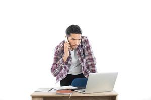 giovane uomo d'affari che parla al telefono mentre lavora al computer portatile in ufficio, tempo occupato con il lavoro isolato su priorità bassa bianca.