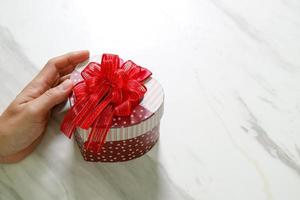 regalo, mano d'uomo che tiene una confezione regalo a forma di cuore in un gesto di dare su sfondo bianco tavolo in marmo grigio foto