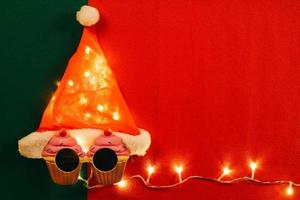 concetto di stagione dei saluti. Cappello di Babbo Natale con luce stellare e occhiali che decorano con cupcake di Natale su sfondo rosso e verde foto