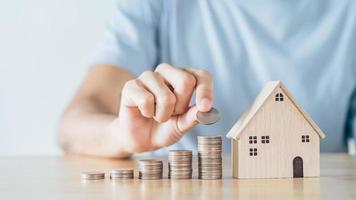 la mano dell'uomo ha messo la moneta sulla pila delle monete con la casa di legno sulla tavola di legno. risparmio di denaro per l'acquisto di casa, concetto di mutuo per la casa del piano finanziario. foto