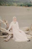 bella modella islamica che indossa la moda hijab, un moderno abito da sposa per donna musulmana seduta sulla sabbia e sulla spiaggia. ritrarre un modello di ragazza asiatica usando l'hijab divertendosi in spiaggia con alberi foto