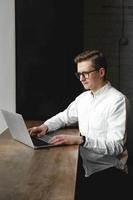 giovane uomo che indossa una camicia bianca e occhiali da vista seduto con il laptop e lavora nella caffetteria foto