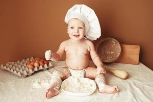 ragazzino panettiere in un cappello da chef seduto sul tavolo che gioca con la farina su uno sfondo marrone con un mattarello in legno, un setaccio rustico rotondo e uova. copia, spazio vuoto per il testo foto