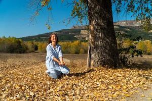 donna sorridente in camicia lunga blu seduta sotto un albero e lanciando foglie autunnali. ritratto femminile autunnale foto