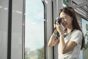 una bella turista asiatica con la macchina fotografica nella cabina passeggeri, che viaggia in treno sopraelevato, scatta foto istantanee durante il trasporto in vista urbana, stile di vita cittadino in treno, felice vacanza di viaggio.