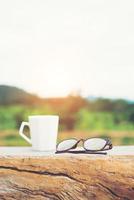 bianco della tazza di caffè caldo con gli occhiali sulla panchina con sfondo verde della montagna della natura. foto