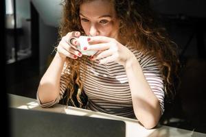 giovane donna caucasica che beve caffè mentre guarda video sul computer portatile a casa al mattino presto foto