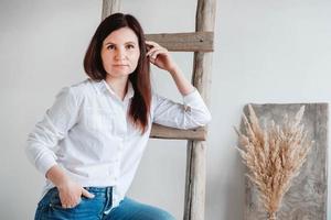 bella donna vestita con una camicia bianca e jeans blu in posa vicino a una scala di legno su sfondo bianco foto