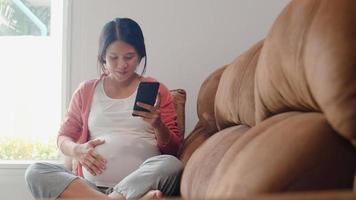 giovane donna incinta asiatica che utilizza il telefono cellulare cerca informazioni sulla gravidanza. la mamma si sente felice sorridente positiva e pacifica mentre si prende cura del suo bambino sdraiato sul divano nel soggiorno a casa concetto. foto