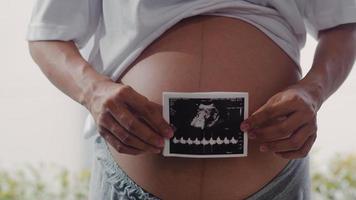 le giovani donne incinte asiatiche mostrano e guardano la foto degli ultrasuoni bambino nella pancia. mamma che si sente felice sorridente tranquillo mentre si prende cura del bambino sdraiato vicino alla finestra nel soggiorno a casa concetto.