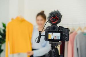 influencer online blogger femminile di moda asiatica con borse della spesa e molti vestiti su appendiabiti per la registrazione di nuovi video di moda trasmessi video in diretta sui social network da Internet a casa. foto