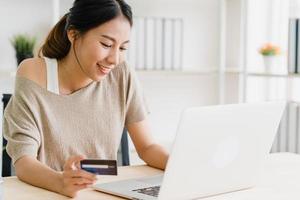 bella donna asiatica che utilizza computer o laptop acquistando acquisti online con carta di credito mentre indossa un maglione seduto sulla scrivania nel soggiorno di casa. donna di stile di vita a casa concetto. foto