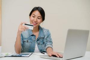 bella donna asiatica che utilizza computer o laptop acquistando acquisti online con carta di credito mentre indossa un maglione seduto sulla scrivania nel soggiorno di casa. donna di stile di vita a casa concetto. foto