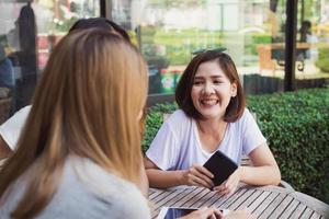 giovani donne asiatiche allegre che si siedono nel caffè che bevono caffè con gli amici e che parlano insieme. donna asiatica attraente che gode del caffè mentre utilizza lo smartphone per parlare, leggere e inviare messaggi di testo. foto