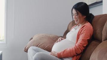 giovane donna incinta asiatica che tiene la sua pancia a parlare con suo figlio. mamma che si sente felice sorridente positiva e pacifica mentre si prende cura del bambino, gravidanza sdraiata sul divano nel soggiorno a casa concetto. foto