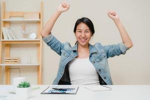 asia donna d'affari celebrazione del successo mantenendo le braccia alzate in ufficio a casa. giovane donna asiatica che lavora scrivendo la finanza del documento del taccuino sulla scrivania in soggiorno a casa. godersi il tempo a casa concetto. foto