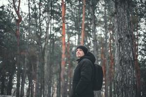 un giovane ragazzo attraente con i capelli corti in un cappotto invernale nero posa sullo sfondo di una foresta invernale. foto